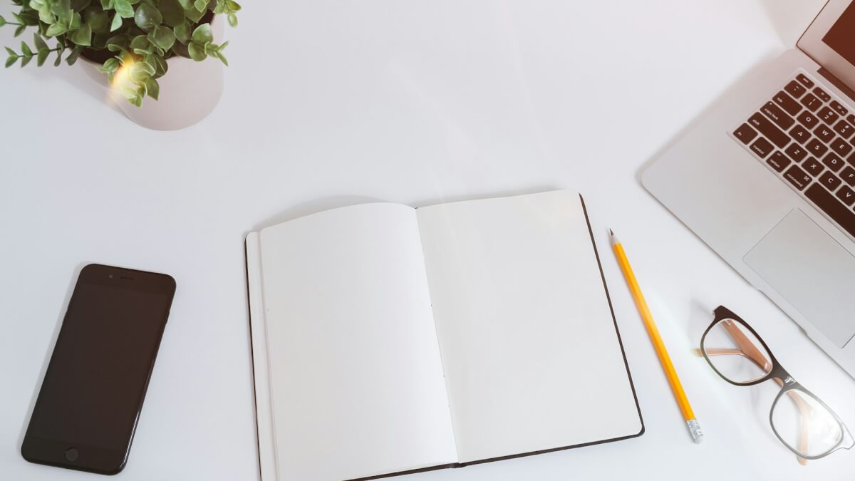 An open empty notebook on a white desk next to an iPhone and a MacBook